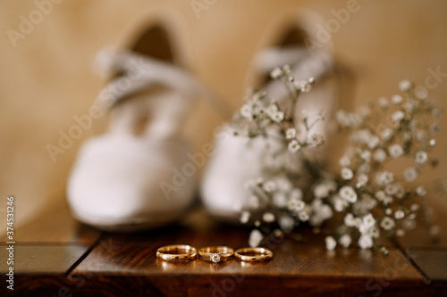 Wedding rings and an engagement ring on a wooden table with womens wedding white shoes and wildflowers.