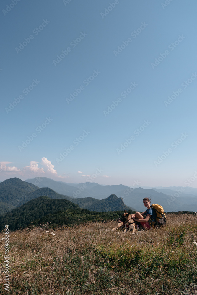 A girl traveler and her German shepherd sit on a hill and look at the mountains around. The dog traveler and its human owner with a backpack walk in the national Park and enjoy nature.