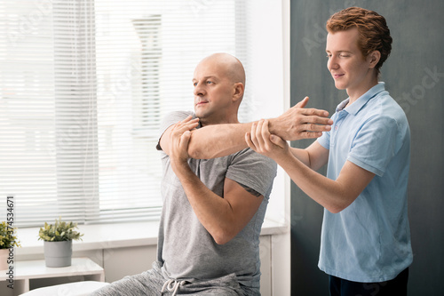 Young contemporary physiotherapist helping mature male patient in clinics