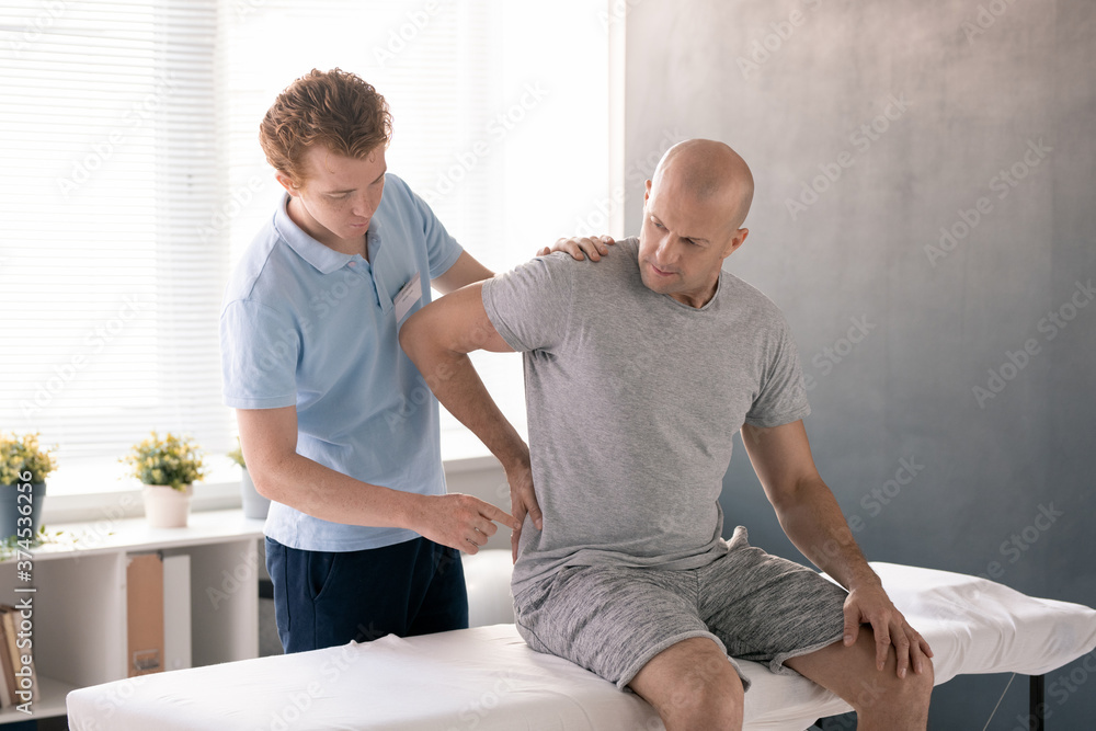 Bald mature man touching painful back while sitting on medical couch in clinics Bald mature man touching painful back while sitting on medical couch in clinics