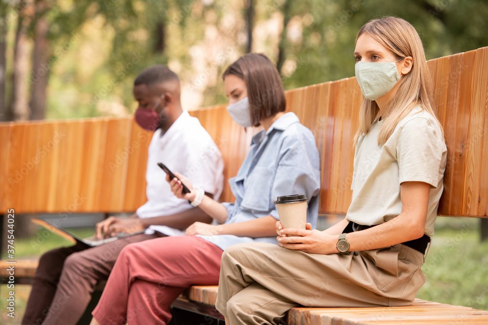 Young blond bored woman in protective mask having coffee while sitting on bench Young blond bored woman in protective mask having coffee while sitting on bench