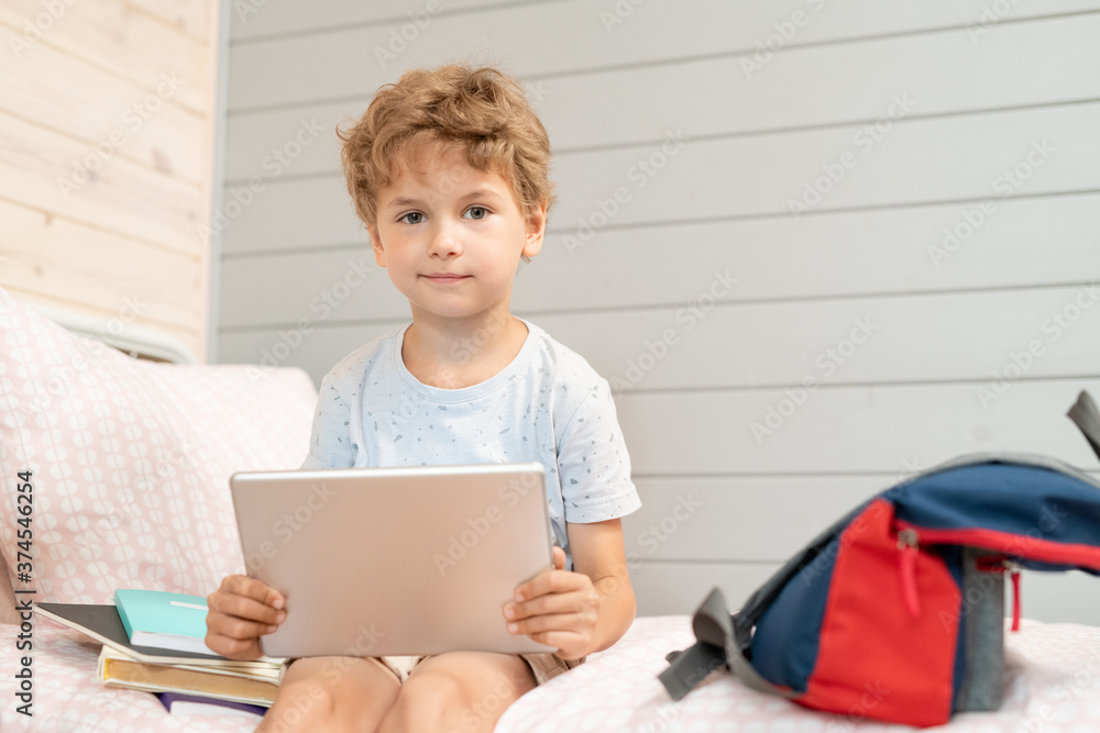Adorable blond schoolboy with digital tablet sitting on bed after school Adorable blond schoolboy with digital tablet sitting on bed after school