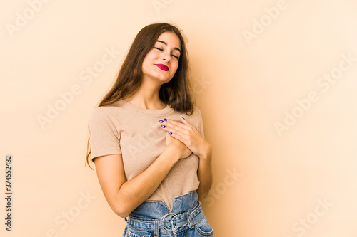 Young caucasian woman isolated en beige background has friendly expression  pressing palm to chest. Love concept.