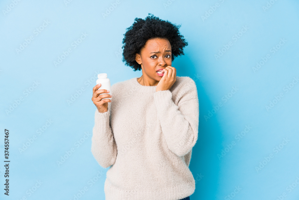 Φωτογραφία Middle age african american woman holding a vitamin bottle biting fingernails, nervous and very anxious