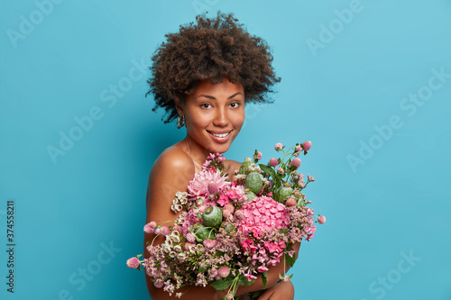Pleasant looking curly woman gets natural holiday gift, carries beautiful bouquet, enjoys mothers day, stands bare shoulders, smiles positively, isolated on blue background, has spring mood.