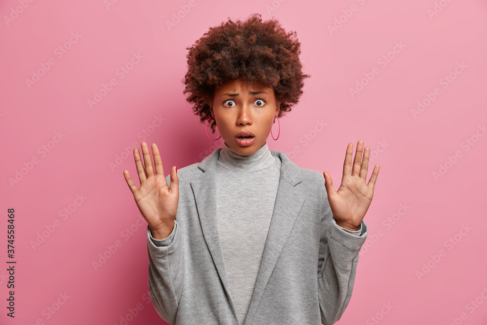 Frightened puzzled dark skinned woman with curly hair raises palms in stop gesture, asks not bother her, dressed in grey formal clothes, has scared expression, isolated on pink studio background. Frightened puzzled dark skinned woman with curly hair raises palms in stop gesture, asks not bother her, dressed in grey formal clothes, has scared expression, isolated on pink studio background.