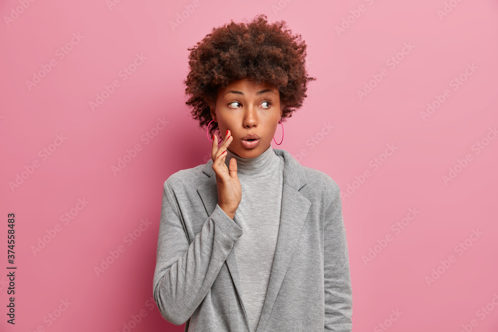 Surprised beautiful curly haired woman with wondered expression, gasps and looks away, being impressed and speechless, wears grey jacket, poses against pink background, sees incredible thing. Surprised beautiful curly haired woman with wondered expression, gasps and looks away, being impressed and speechless, wears grey jacket, poses against pink background, sees incredible thing.