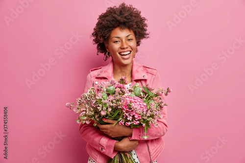 Isolated shot of carefree joyful dark skinned woman poses with mix of field flowers, embraces big bouquet, dressed in stylish clothes, enjoys blooming gift. Mothers Day, celebration concept.