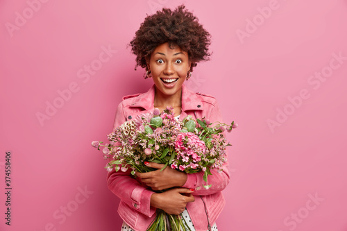 Amused surprised dark skinned woman embraces bouquet of pretty flowers, going to congratulate friend with anniversary, wears pink fashionable jacket, stands indoor. Celebration, special occasion