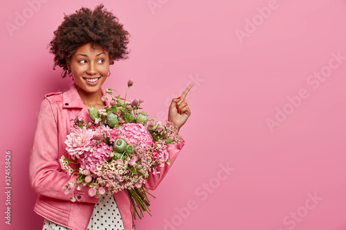 Romantic positive young woman with Afro hair points index finger to side, holds pretty bouquet of mix flowers, shows copy space for your advertisement, rosy background, gives way to flower shop