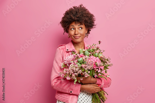 Pensive Afro American woman thinks about celebration and party holiday, receieves beautiful bouquet on Valentines day, dressed in fashionable clothes, isolated on pink wall. Flower delivery concept