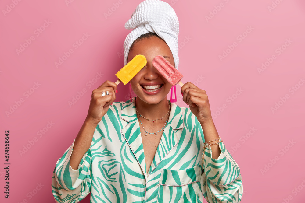 Summer is almost over. Positive dark skinned woman covers eyes with fresh cold ice creams, has fun during hot day, wears casual domestic robe and bath towel after taking shower, rosy background Summer is almost over. Positive dark skinned woman covers eyes with fresh cold ice creams, has fun during hot day, wears casual domestic robe and bath towel after taking shower, rosy background