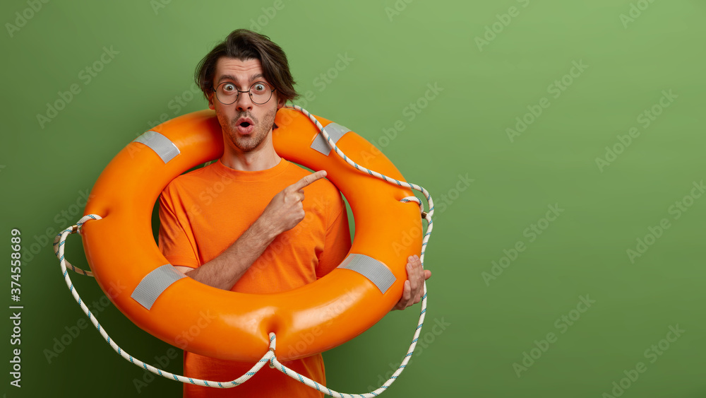 © wayhome.studio  - Emotional stunned man poses with safety equipment, uses lifebuoy on rescue, points at copy space, has shocked expression, isolated over green background. Time for swimming. Accident prevention