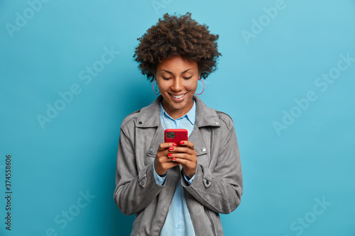Happy smiling curly haired young woman types message on mobile phone, looks with glad expression at display, wears grey jacket, isolated on blue background. People, lifestyle, technology concept