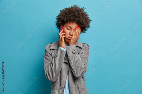 Tired sleepy woman with Afro hair makes face palm, feels tired and fatigue, has boring telephone conversation, calls friend, wears grey jacket, poses against blue background, has red manicure.
