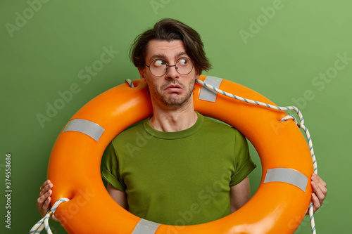 Surprised man poses with buoy ring around neck, looks suspiciously aside, learns to swim, wears transparent glasses and green t shirt, being rescued from water, isolated over green studio wall