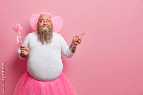 Foto Funny man wears fairy costume, invites you on holiday or costume party, indicates right at blank space, holds magic wand, poses against rosy wall