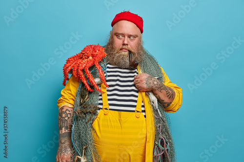 Male mariner looks with scared expression at octopus, afraids big sea creature, poses with fishing net, smokes pipe, dressed in yellow overalls poses indoor. Fishering, marine life, boat trips concept