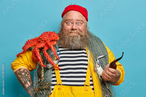 Wallpaper Mural Photo of bearded experienced sailor poses with fishing net, big red crab on shoulders, smoke pipe, welcomes on board, enjoys marine cruise during summer vacation