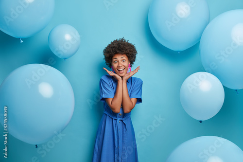 Smiling Afro American girl spreads palms over face, enjoys awesome summer party, poses over inflated balloons in long blue fashionable dress, being in happy mood. Celebration and lifestyle concept