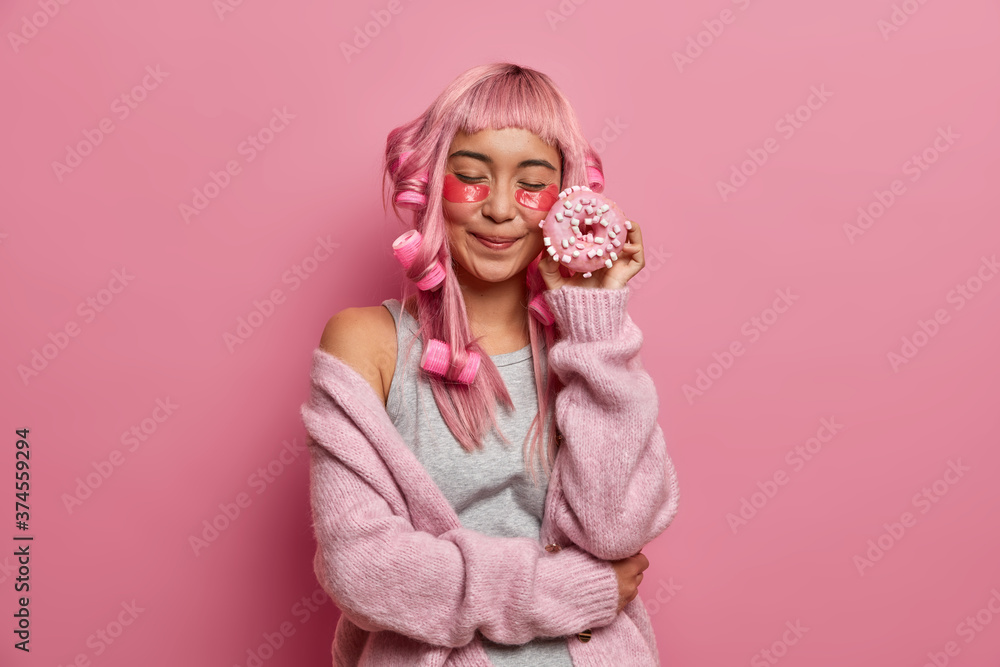 Pleased young Asian woman with pink hair, closes eyes holds delicious doughnuts near face, applies collagen patches under eyes, makes curly hairstyle, wears warm pink jumper, prepares to party Pleased young Asian woman with pink hair, closes eyes holds delicious doughnuts near face, applies collagen patches under eyes, makes curly hairstyle, wears warm pink jumper, prepares to party