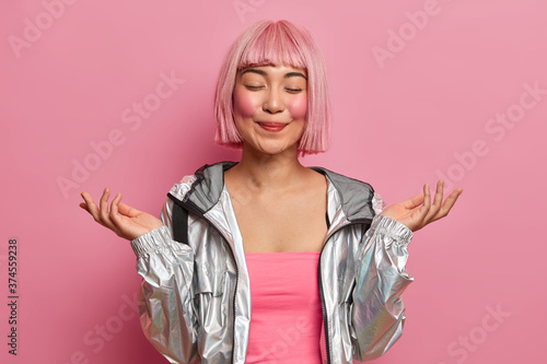 Vászonkép Half length shot of pleased Asian woman raises hands, stands with eyes closed, feels happy, imagines something pleasant, wears silver jacket isolated on pink background