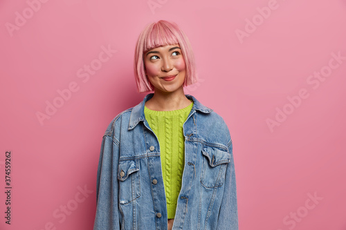 Foto Pleased lovely young Asian girl with dreamy expression, romantic mood, looks somewhere above, wears knitted green jumper and denim jacket, has bob hairstyle, isolated on pink studio background