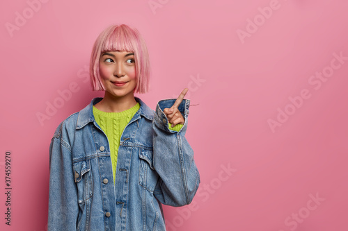 Pleased rouge Asian girl with bob hair, dressed in oversized denim jacket, points on copy space, advertises place, isolated over pink backgound. Eastern woman promoter advertises good product.