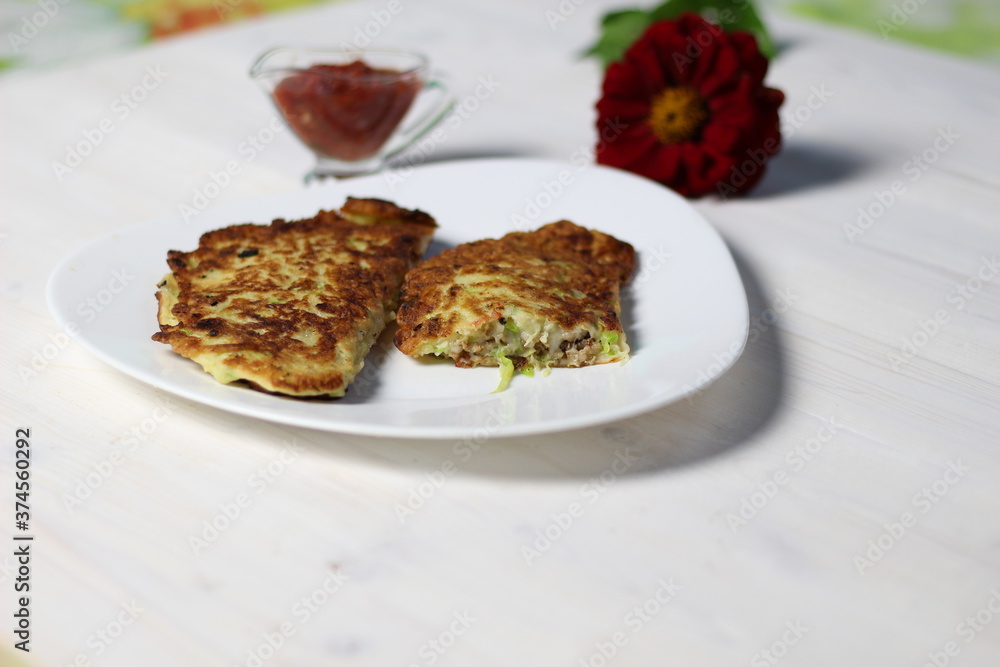 Fried squash pancakes with meat mince and cheese on white plate with tomato sauce and red flower on wooden table Fried squash pancakes with meat mince and cheese on white plate with tomato sauce and red flower on wooden table