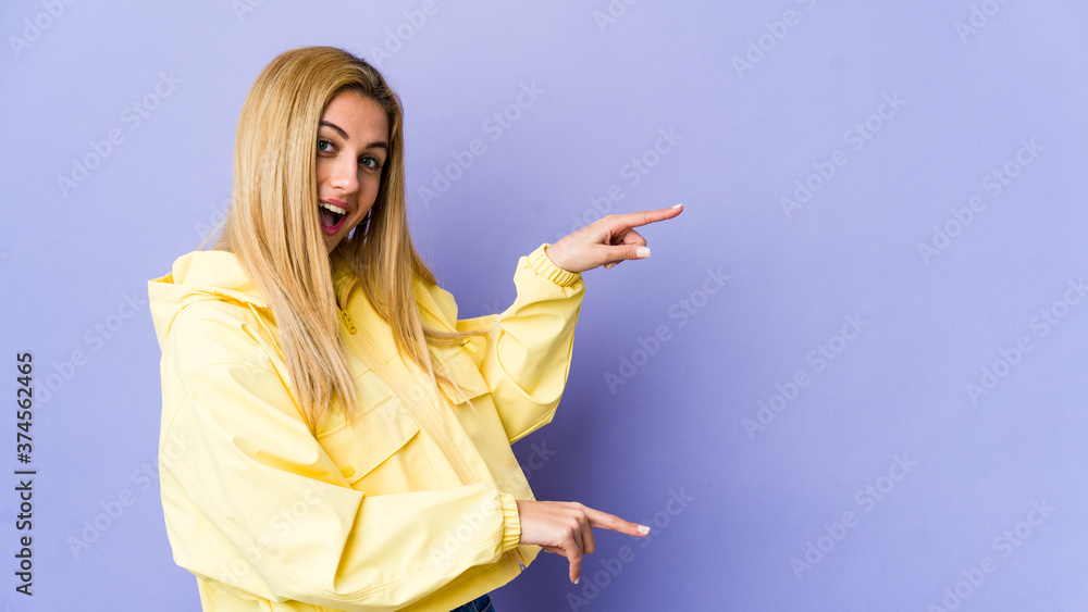 Young blonde woman isolated on purple background excited pointing with forefingers away. Young blonde woman isolated on purple background excited pointing with forefingers away.