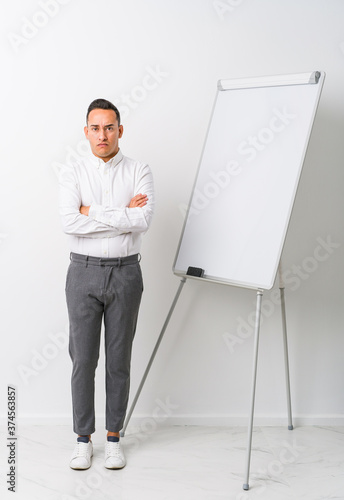 Young latin coaching man with a whiteboard isolated frowning face in displeasure, keeps arms folded.