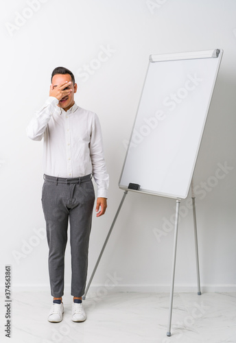 Young latin coaching man with a whiteboard isolated blink at the camera through fingers, embarrassed covering face.