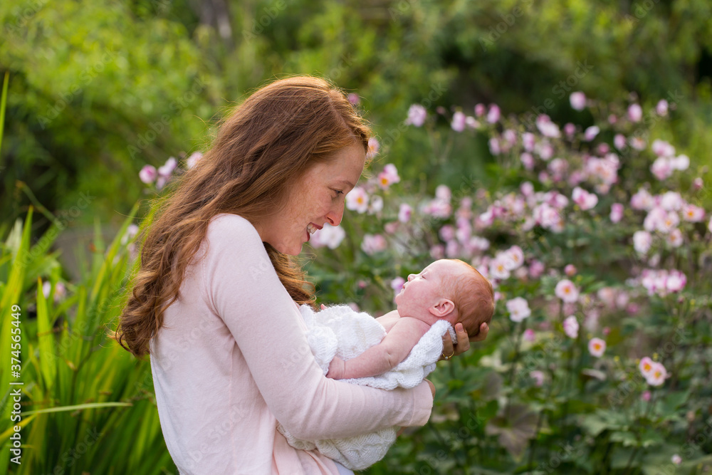 Medium horizontal profile shot of pretty red-haired young woman holding and smiling at her newborn baby girl tenderly while standing in garden Medium horizontal profile shot of pretty red-haired young woman holding and smiling at her newborn baby girl tenderly while standing in garden