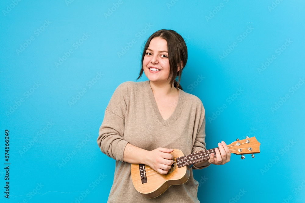 Young caucasian curvy woman playing ukelele isolated on a blue background Young caucasian curvy woman playing ukelele isolated on a blue background