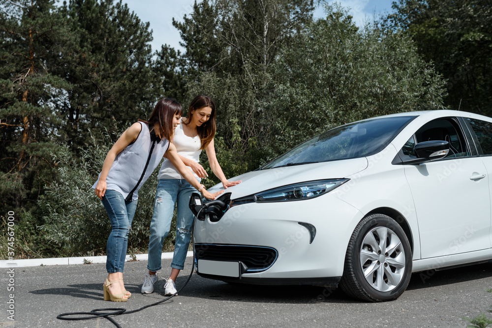 Two happy young beautiful women are talking to each other until their electric car is charging at the charging station situated in the forest. Two happy young beautiful women are talking to each other until their electric car is charging at the charging station situated in the forest.