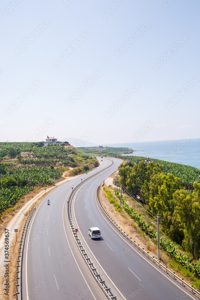An empty asphalt road winds along the breathtaking scenic coastline on a sunny summer day. A spectacular shot of a coastal road overlooking clear blue skies and the calm Mediterranean Sea. An empty asphalt road winds along the breathtaking scenic coastline on a sunny summer day. A spectacular shot of a coastal road overlooking clear blue skies and the calm Mediterranean Sea.