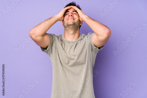 Young man isolated on purple background laughs joyfully keeping hands on head. Happiness concept.