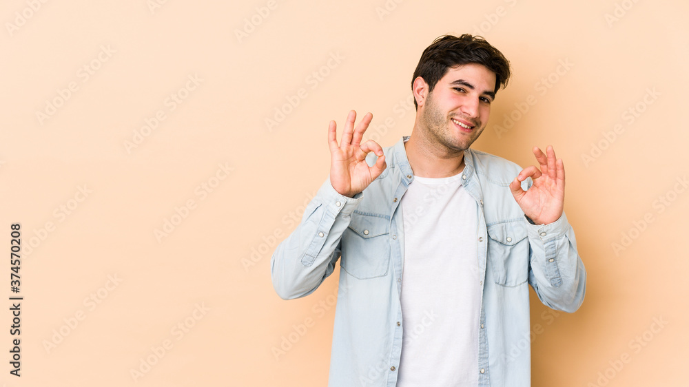 Young man isolated on beige background cheerful and confident showing ok gesture. Young man isolated on beige background cheerful and confident showing ok gesture.