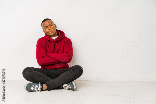 Young latin man sitting on the floor isolated frowning face in displeasure, keeps arms folded.
