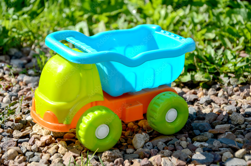 Children's plastic multi-colored toy dump truck standing on the gravel against the background of a green lawn on a sunny lazy day. Toy vehicles, outdoor games for children. Children's plastic multi-colored toy dump truck standing on the gravel against the background of a green lawn on a sunny lazy day. Toy vehicles, outdoor games for children.