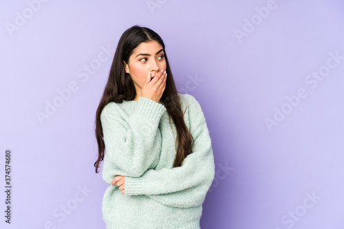 Young indian woman isolated on purple background thoughtful looking to a copy space covering mouth with hand. © Asier
