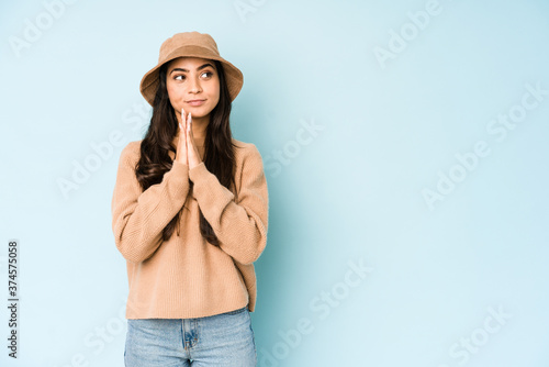 Young indian woman wearing a hat isolated on blue background praying, showing devotion, religious person looking for divine inspiration.