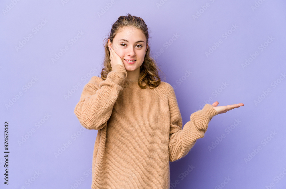 Young caucasian woman on purple background holds copy space on a palm, keep hand over cheek. Amazed and delighted. Young caucasian woman on purple background holds copy space on a palm, keep hand over cheek. Amazed and delighted.