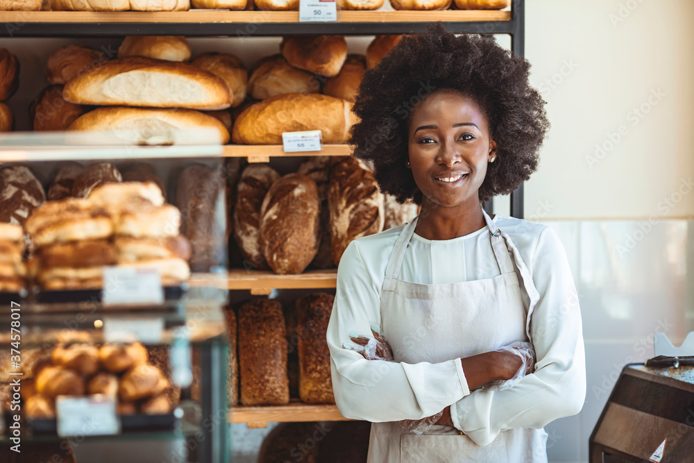 Portrait of positive African American young woman working in own Bakery shop, looking at camera with toothy smile. Pretty baker smiling at camera. Small Bakery shop owner standing in front of store - obrazy, fototapety, plakaty 