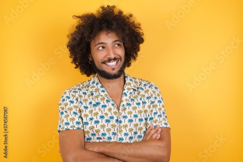 Dreamy rest relaxedYoung man with afro hair over wearing hawaiian shirt standing over yellow background crossing arms, looks good © Roquillo