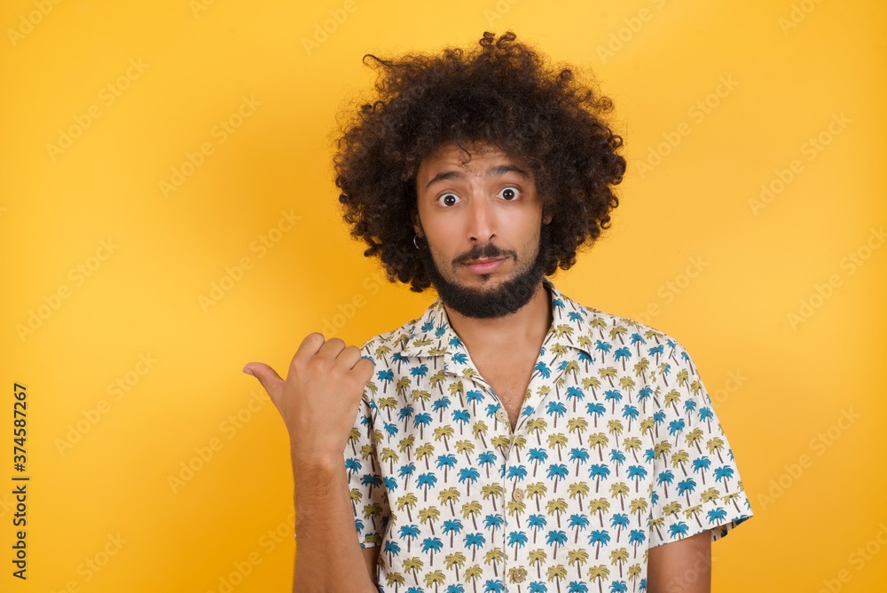 Lovely Young man with afro hair over wearing hawaiian shirt standing over yellow background pointing aside with forefinger, showing at copy space having news about bargains Lovely Young man with afro hair over wearing hawaiian shirt standing over yellow background pointing aside with forefinger, showing at copy space having news about bargains