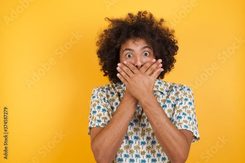 Young man with afro hair over wearing hawaiian shirt standing over yellow background shocked covering mouth with hands for mistake. Secret concept.
