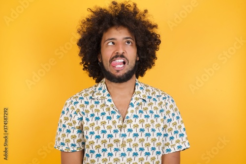 Young man with afro hair over wearing hawaiian shirt standing over yellow background showing grimace face crossing her eyes and showing tongue . Being funny and crazy