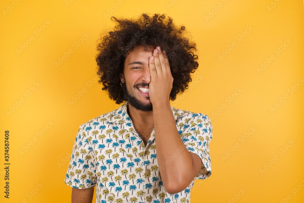 Φωτογραφία Young man with afro hair over wearing hawaiian shirt standing over yellow background covering one eye with her hand, confident smile on face and surprise emotion