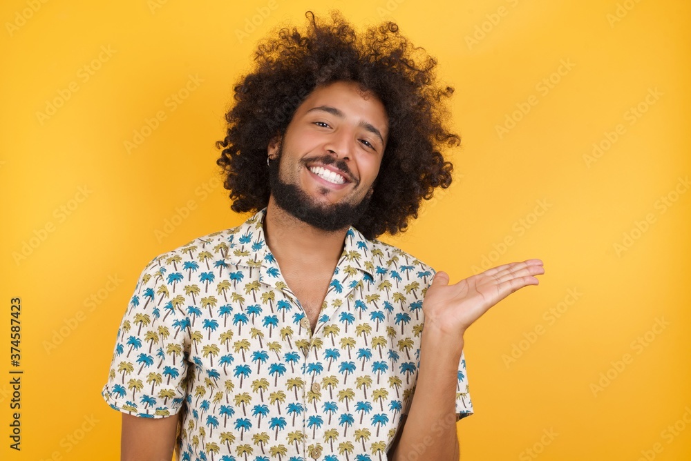 Φωτογραφία Young man with afro hair over wearing hawaiian shirt standing over yellow background smiling cheerful presenting and pointing with palm of hand looking at the camera